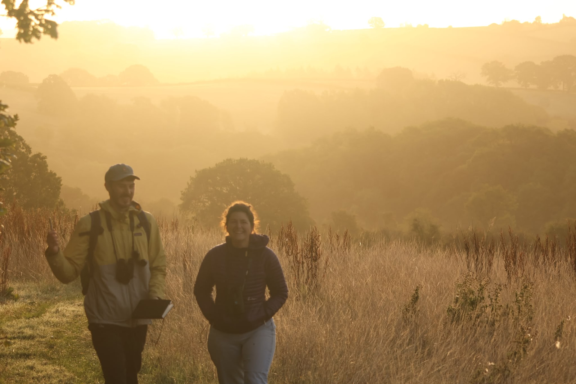 Two people walking through long grass at sunrise